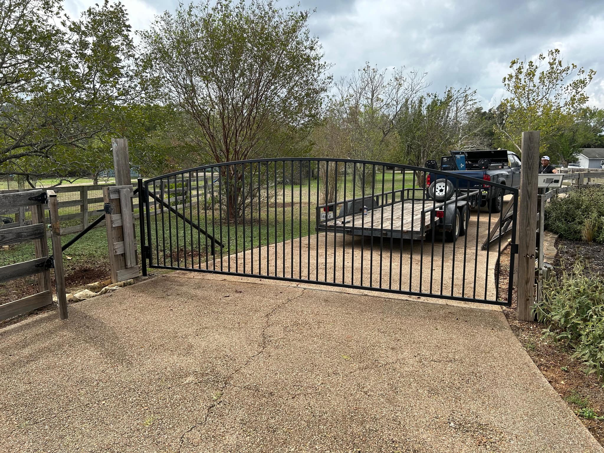 Arched black iron driveway gate hung between cedar posts in a wood rail fence