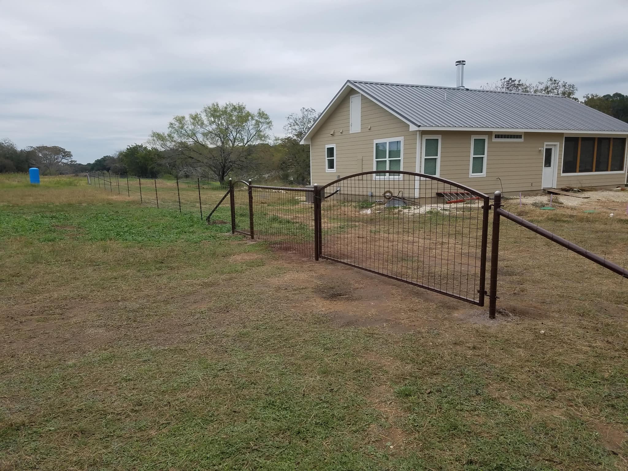 Arched brown steel gate with wire panel fill in a field fence, in front of a house