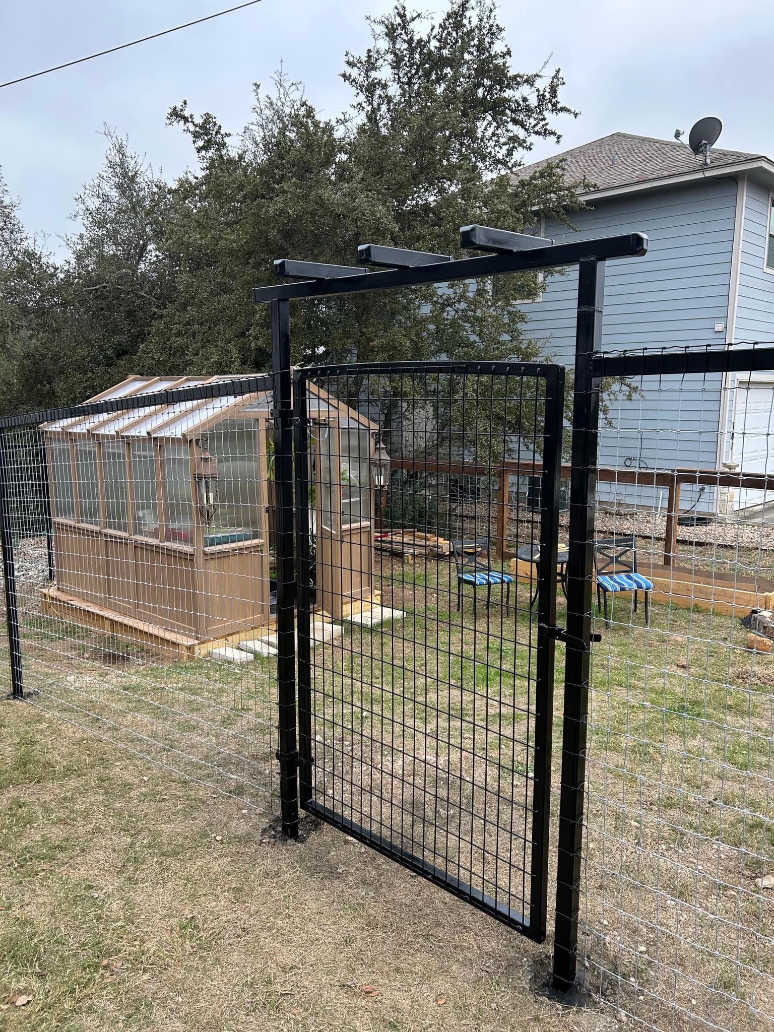 Black wire panel garden fence with an arbor-topped gate around a greenhouse