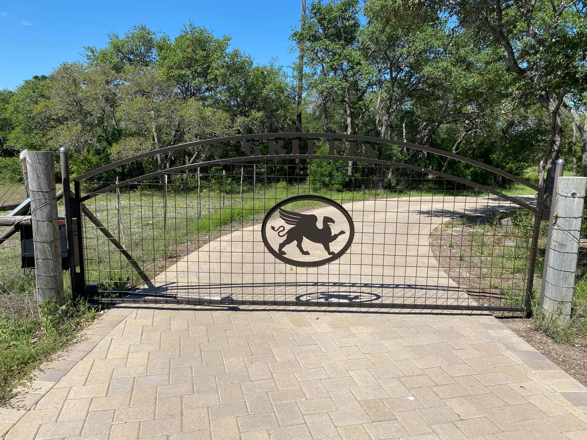 Arched wire-panel driveway gate with a cut-steel griffin medallion over a paver drive