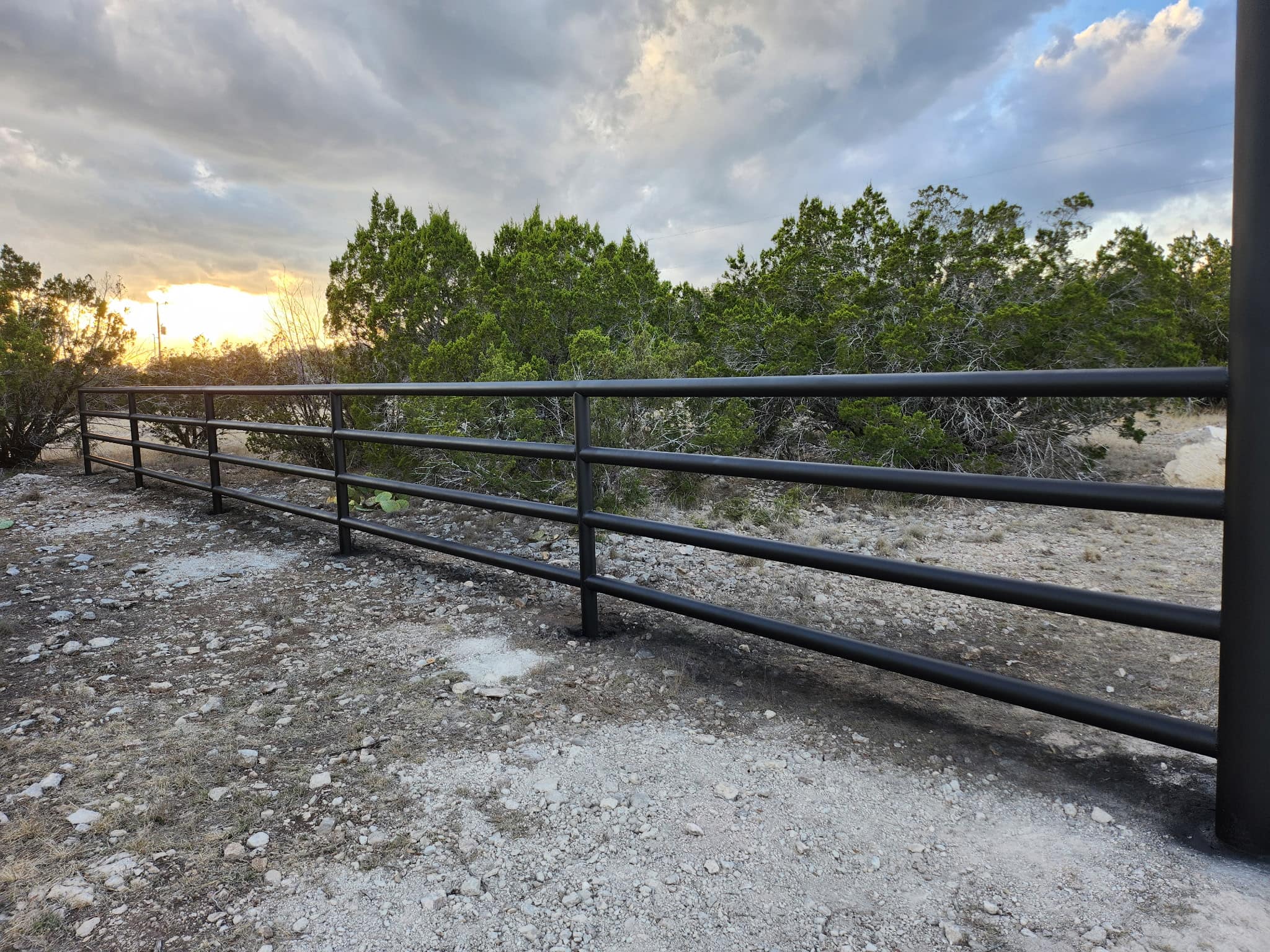 Black four-rail steel pipe fence on rocky ground at sunset