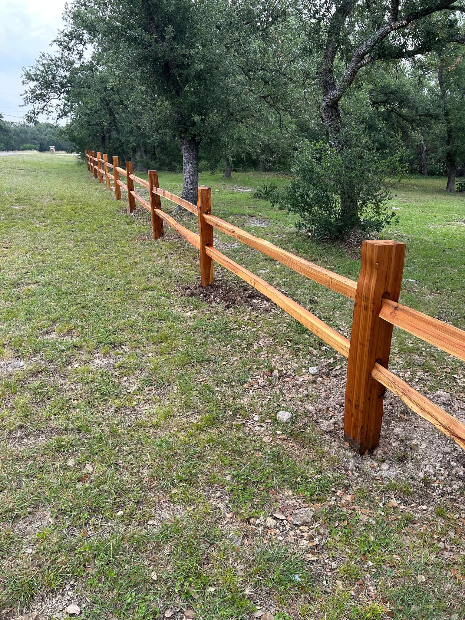 Stained wood two-rail post and rail fence along a pasture under oaks
