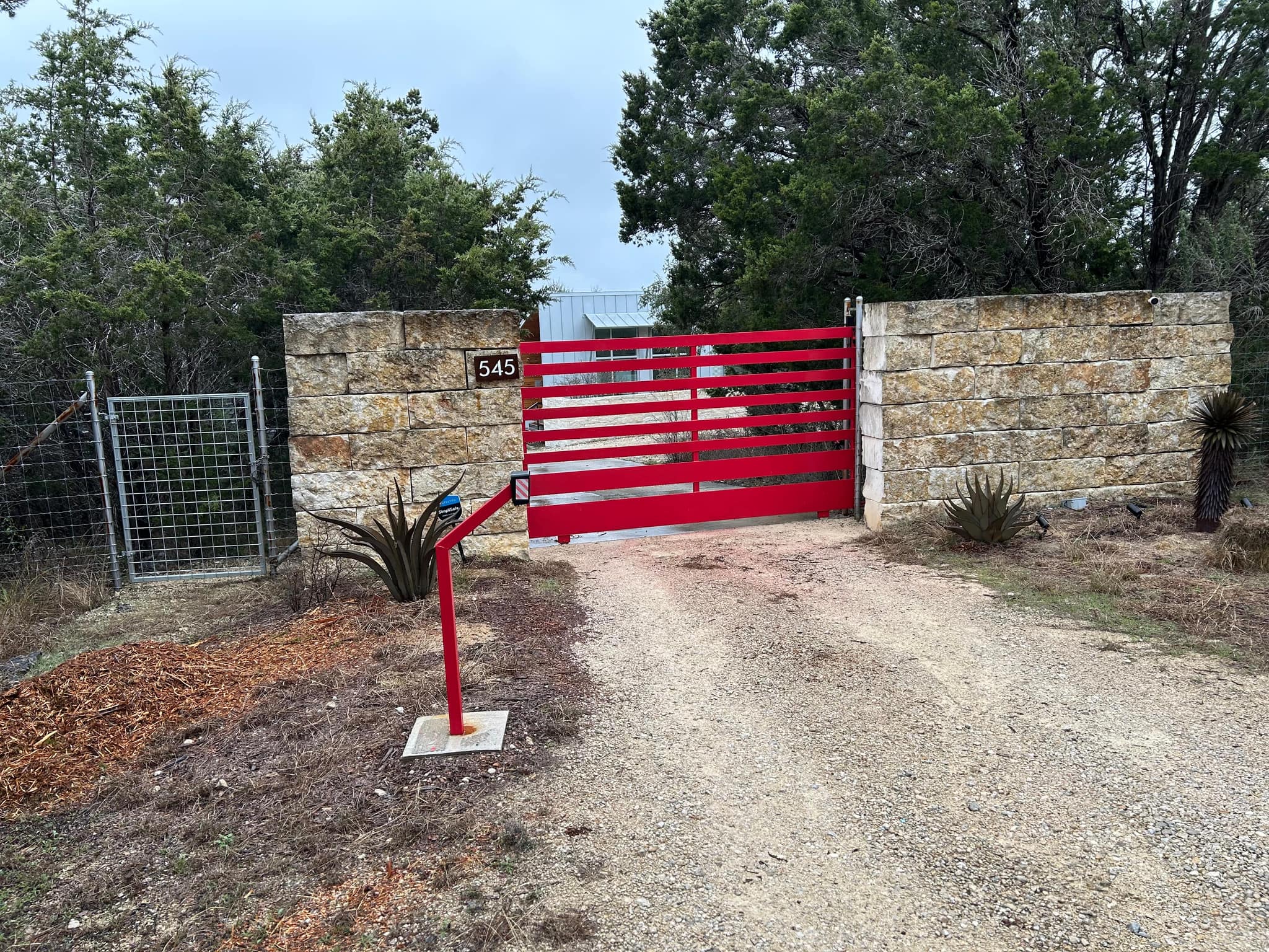 Red horizontal-slat driveway gate between stone walls, with a keypad post and a wire walk gate