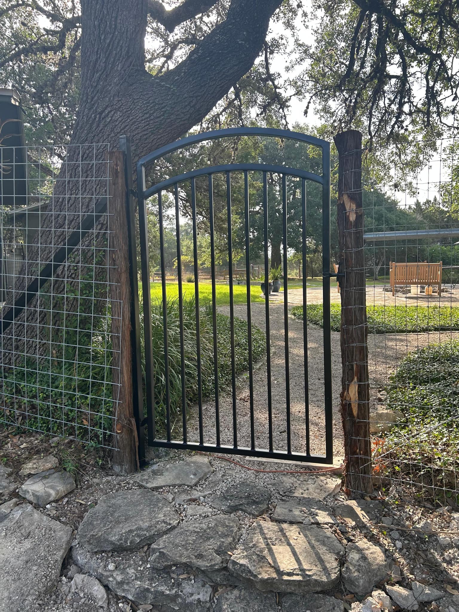 Arched black iron walk gate hung on cedar posts in a welded-wire fence